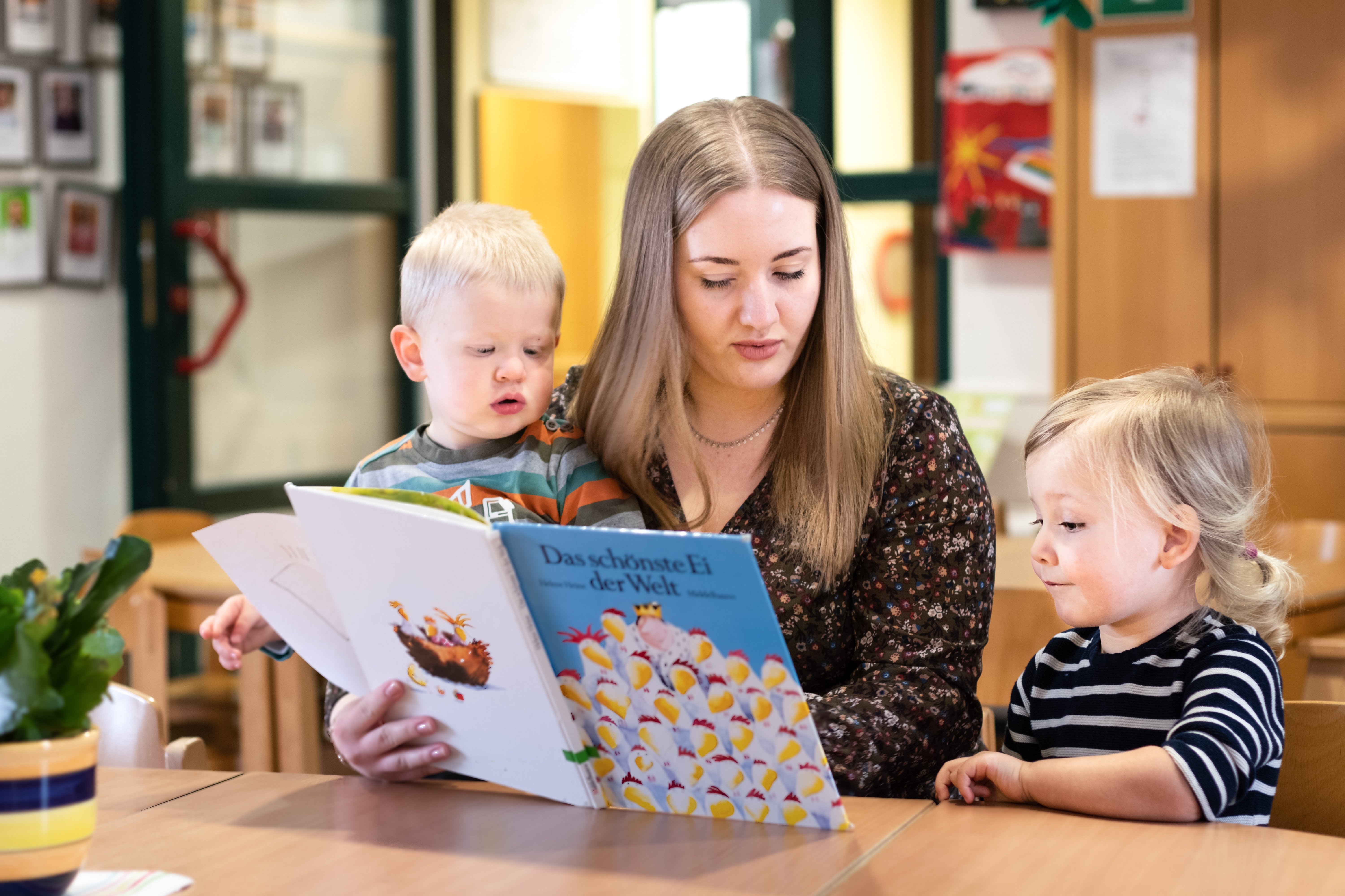 Eine junge Frau liest mit zwei Kleinkindern ein Buch. Sie sitzen an einem Tisch.