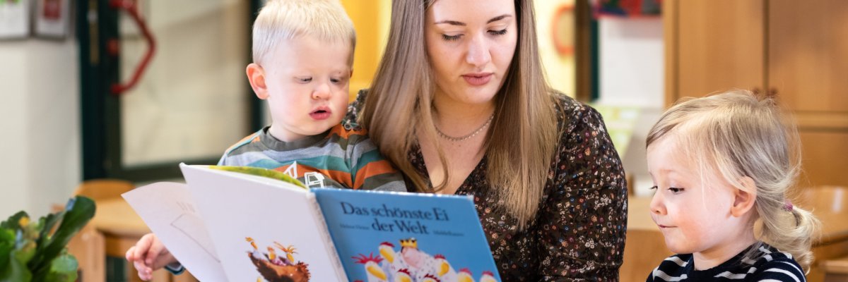 Symbolbild Erzieherin Eine junge Frau liest mit zwei Kleinkindern ein Buch. Sie sitzen an einem Tisch.