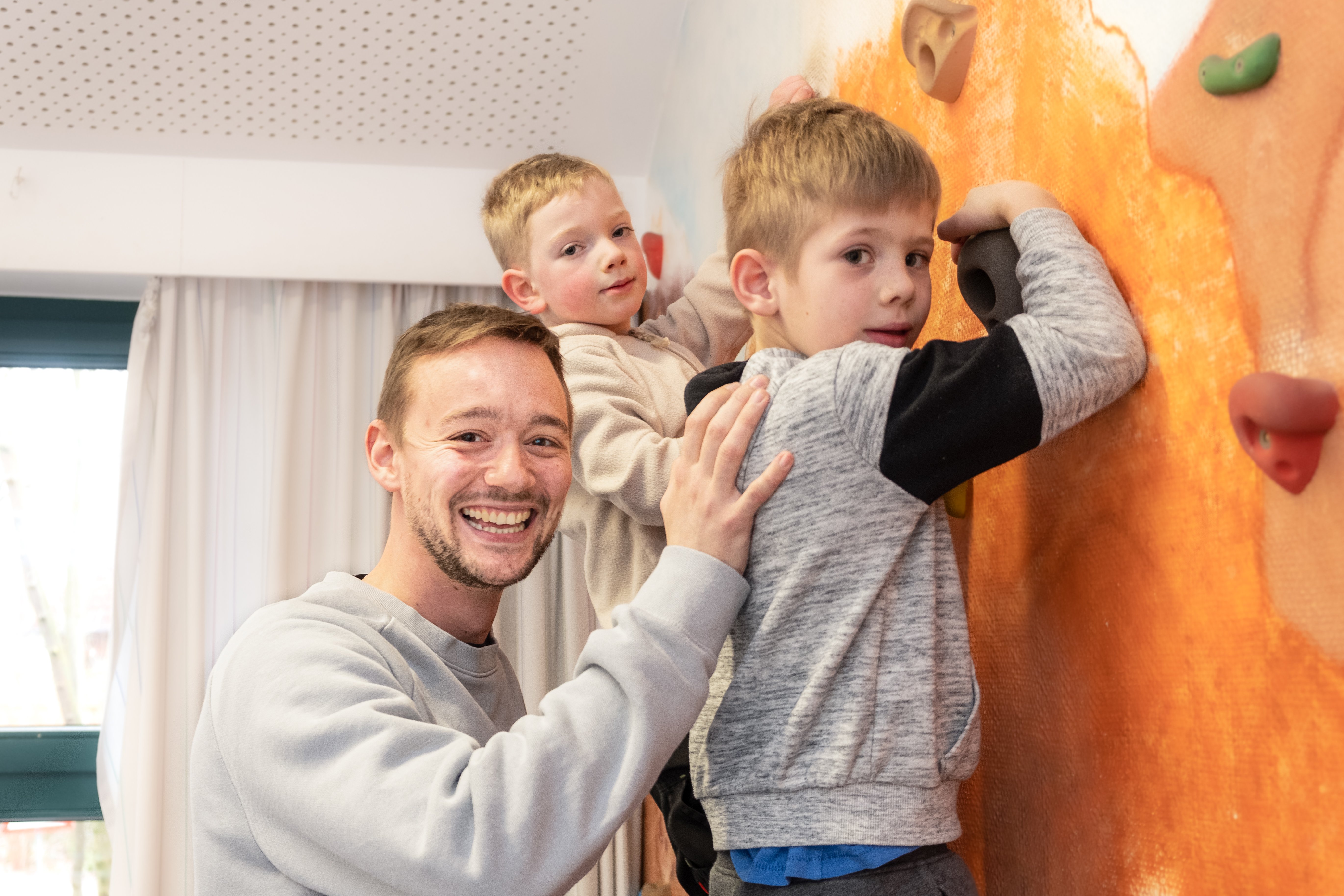 Ein junger Mann unterstützt zwei Kinder an der Kletterwand, alle drei Personen schauen und die Kamera und lächeln.