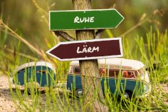 Zwei Wegweiser auf einer Wiese mit einem VW Bus. Das obere Schild ist grün, zeigt nach rechts mit der Aufschrift Ruhe. Das untere Schild ist rot, zeigt nach links mit der Aufschrift Lärm.