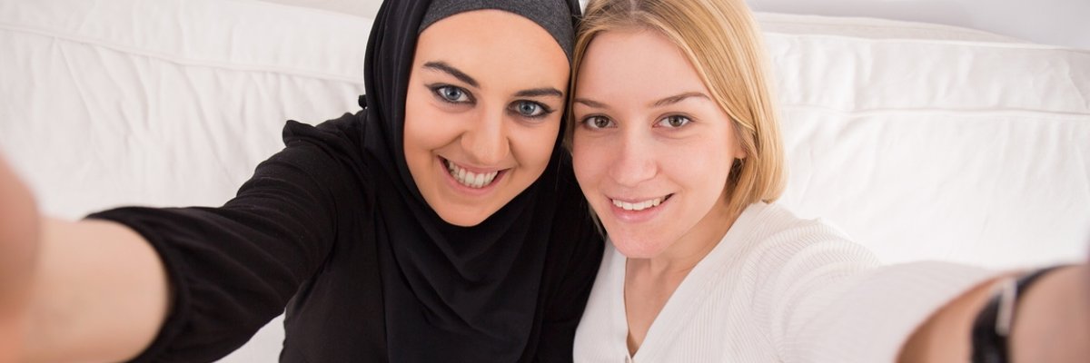 Zwei junge Frauen halten sich im Arm und lächeln in die Kamera. Eine der beiden Frauen trägt ein Kopftuch. 