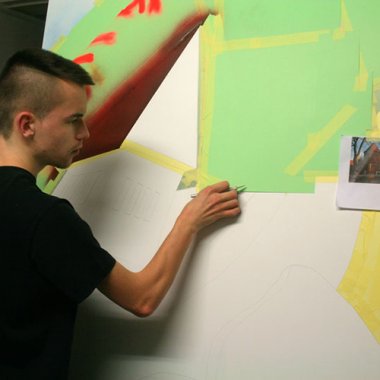 Künstler Büren Ein junger Mann klebt auf einer weißen Holzplatte einzele Parzellen mit Klebeband ab, erste Bereiche sind schon grün angemalt.