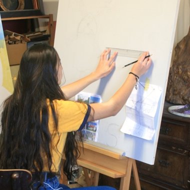 Künstlerin Dudensen Eine junge Frau wird von hinten gezeigt, wie sie vor einer weißen Holzplatte steht und mit einem Lineal Striche einzeichnet.