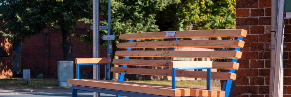 Mitfahrbank Holzbank mit blauem Gestellt steht vor enem Gebäude.