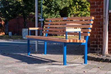 Mitfahrbank Holzbank mit blauem Gestellt steht vor enem Gebäude.