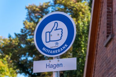 Hinweisschild Mitfahrbank Hagen Rundes Schild, weißer Hintergrund mit blauem Kreis. Darin ein Piktogramm mit Daumen hoch. Darunter ein kleineres rechteckiges Schild mit der Aufschrift Hagen