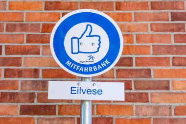 Hinweisschild Mitfahrbank Eilvese Rundes Schild, weißer Hintergrund mit blauem Kreis. Darin ein Piktogramm mit Daumen hoch. Darunter ein kleineres rechteckiges Schild mit der Aufschrift Eilvese