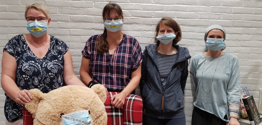 Vier Kolleginnen der Stadtbibliothek mit Masken und Aufschrift "Wir sind wieder da"