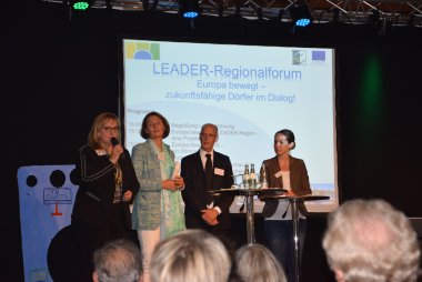 Auf einer Bühne stehen vier Personen. Im Hintergrund steht eine Leinwand. Es geht um die Veranstaltung Leader-Regionalforum