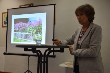 Frau Bukies steht an einem Tisch. Im Hintergrund ist eine Leinwand mit einem Foto von Blumen zu sehen.