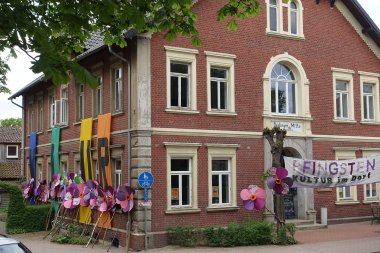 Im Vordergrund eines Gebäudes sind große selbstgebastelte Blumen auf Stöcken aufgestellt. Vor dem Gebäude ist ein großer Banner mit der Aufschrift - Pfingsten Kultur im Dorf -