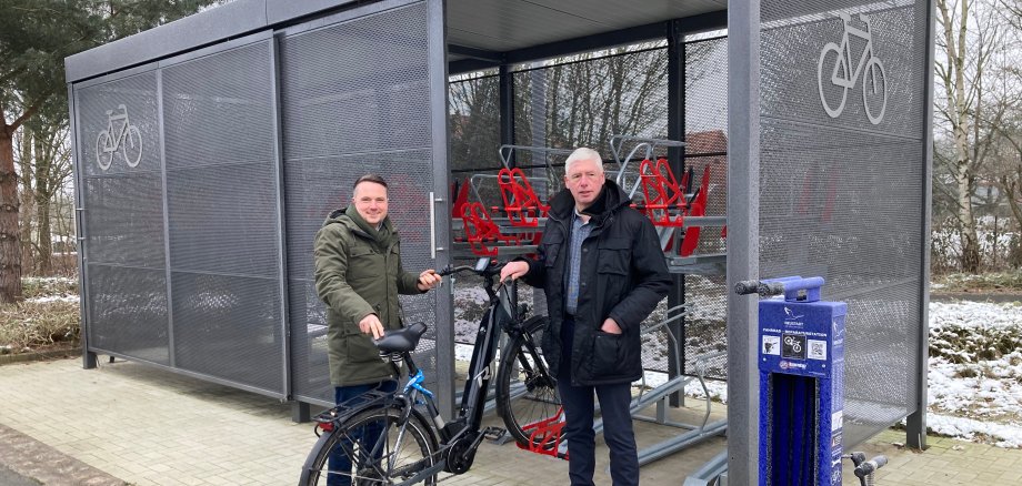 Zwei Herren stehen mit einem Fahrrad an einer Sammelschließanlage für Fahrräder. Das Fahrrad ist mit dem Vorderreifen in der halterung. Die Schließanlage ist ein großer grauer Metallkasen, wi auf zwei ebenen halterungen zum Abstellen von Fahrrädern angebracht sins.