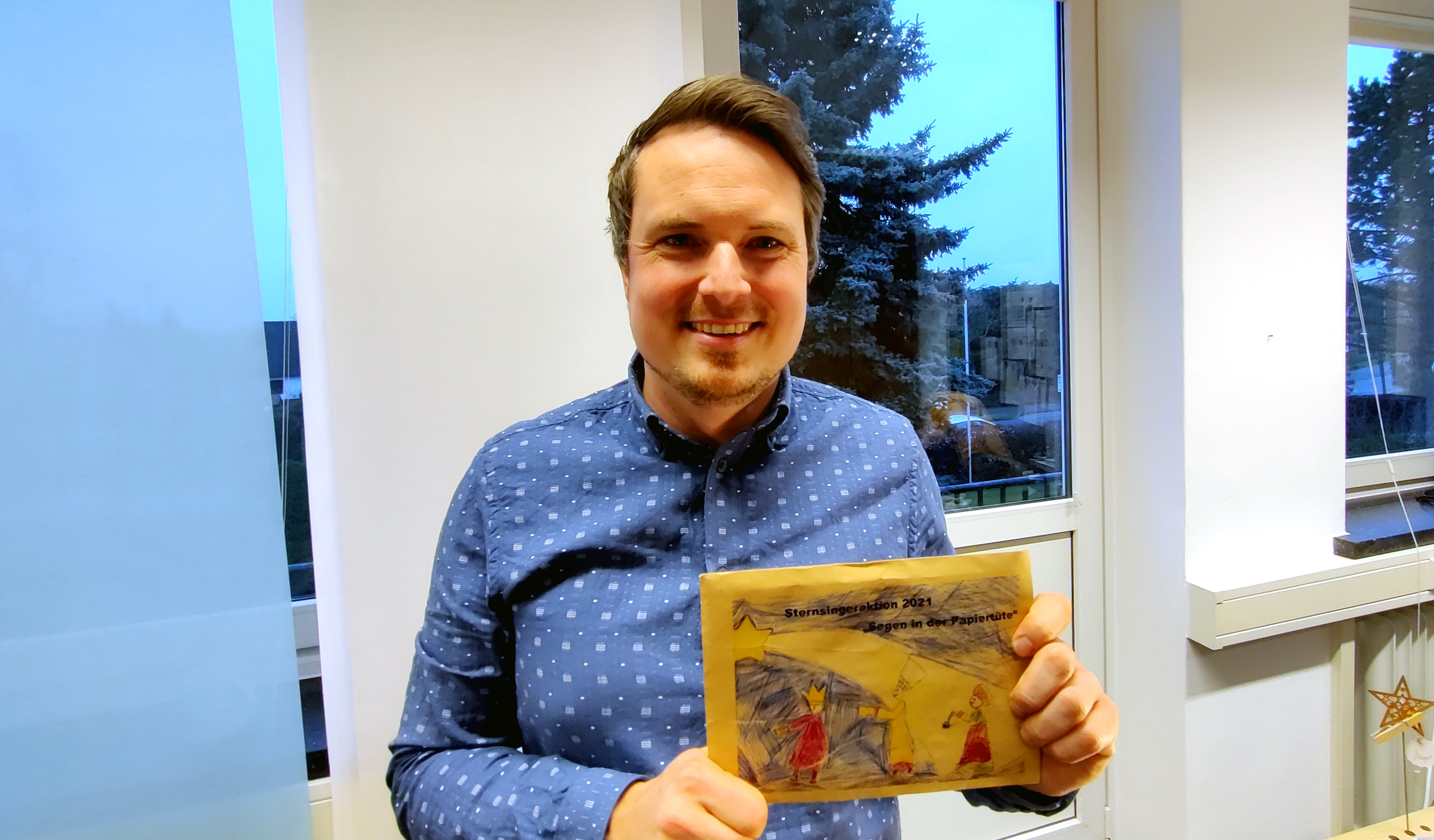 Bürgermeister Dominic Herbst hält einen bemalten Briefumschlag in die Kamera. Darauf steht Sternsingeraktion 2021 "Segen in der Papiertüte"