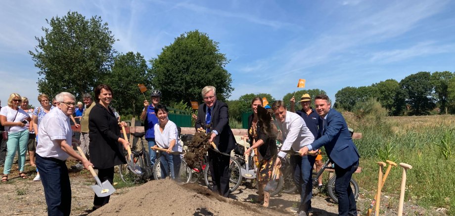 7 Personen stehen hinter einem Erdhügel und werfen gleichzeitig mit Spaten Erde in die Luft. Symbolischer Spatenstich.