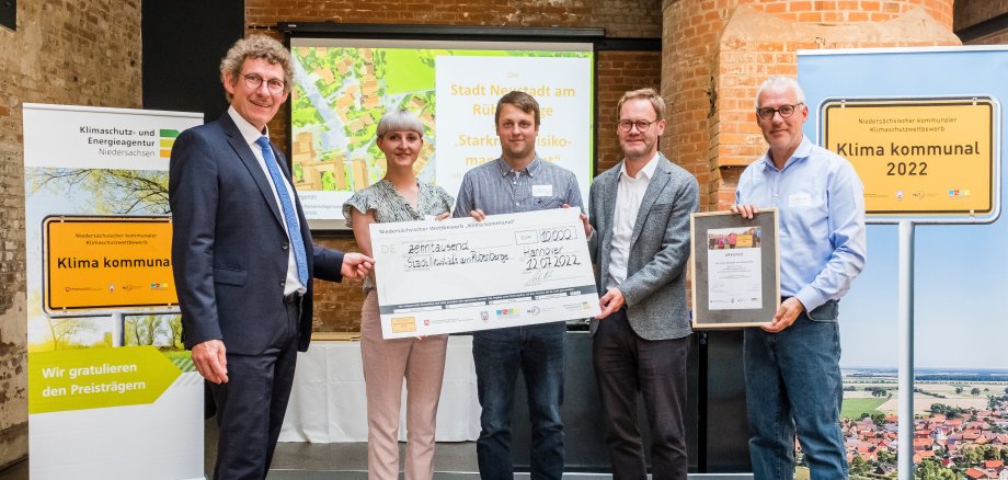 Preisverleihung Starkregenrisikomanagement 5 Personen stehen auf einer Bühne, die mittleren drei sind VertreterInnen der Stadt Neustadt, sie halten einen großen symbolischen Scheck in den Händen. Alle schauen in die Kamera.