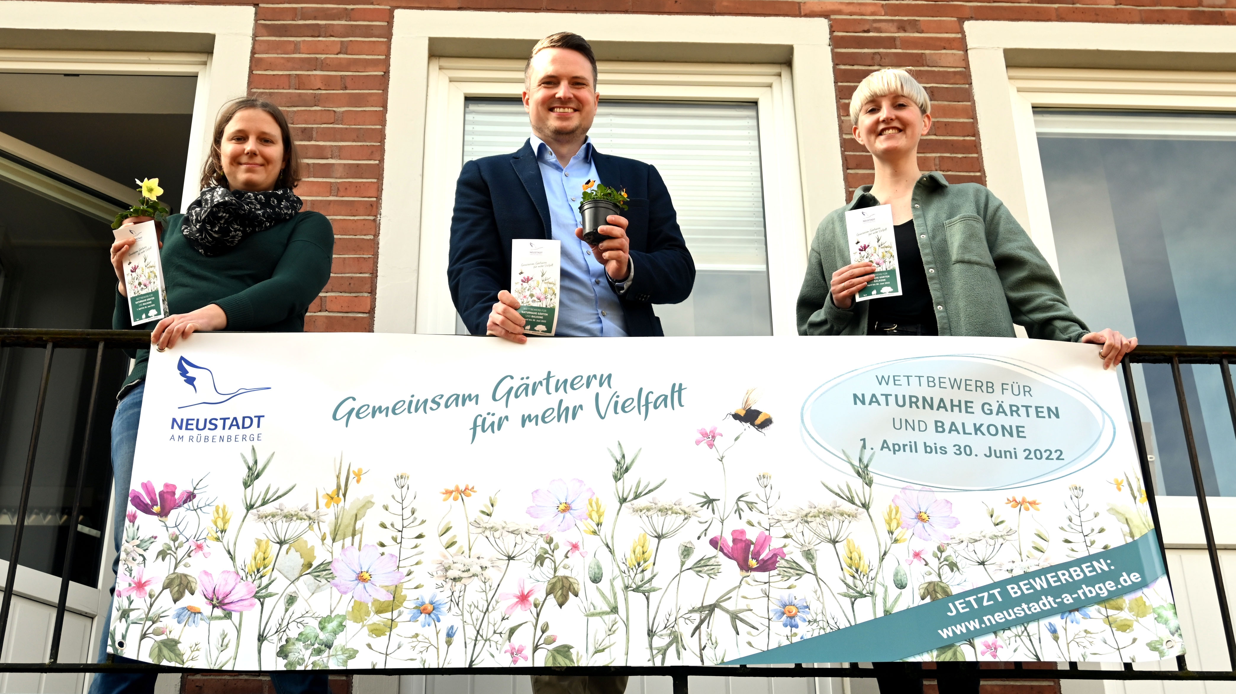 Drei personen stehen auf dem Balkon des Rathauses und halten Blumen und Samentüten in die Kamera. Am Balkongitter hängt ein Banner für den Garten- und Balkonwettbewerb.