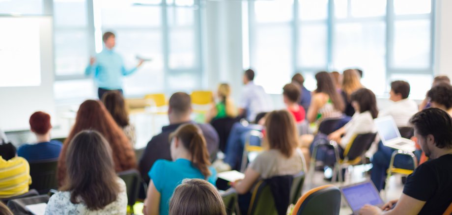 Symbolbild Workshop Eine Gruppe von Menschen sitzt in einem Seminarraum. Im Hintergrund hält eine Person einen Vortrag.