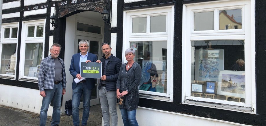 StartPlatz Windmühlenstr. 6 vier Personen stehen vor einem Fachwerkhaus in der Windmühlenstr. 6. Die beiden Herren in der Mitte halten ein Schild hoch, auf dem Start Platz zu lesen ist.