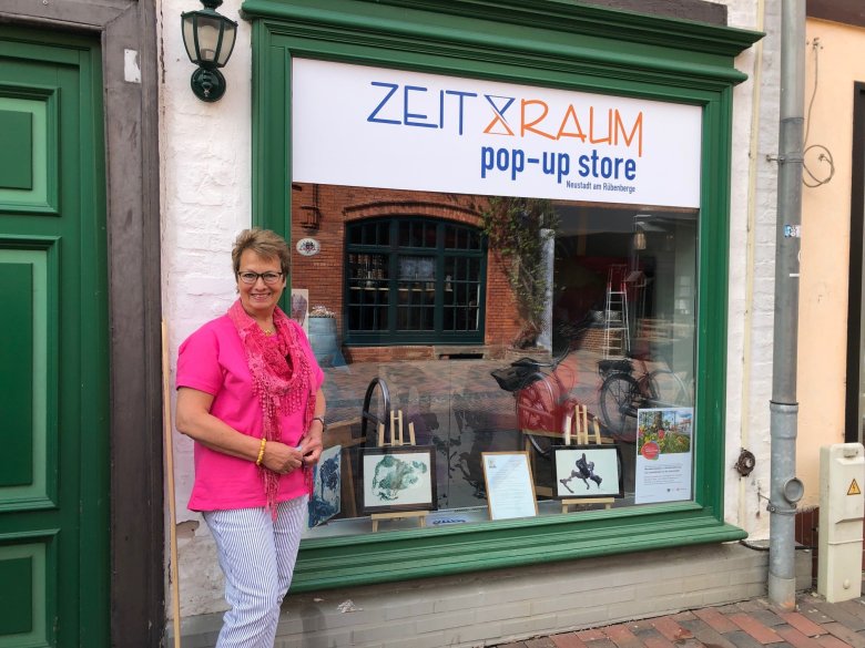 ZeitRaum Markststr. 41 Eine Frau steht links vor einem Schausvenster eines Fachwerkgebäudes in der Markstr. 41. Im Schaufenster sind verschiedene Malerein zu sehen.