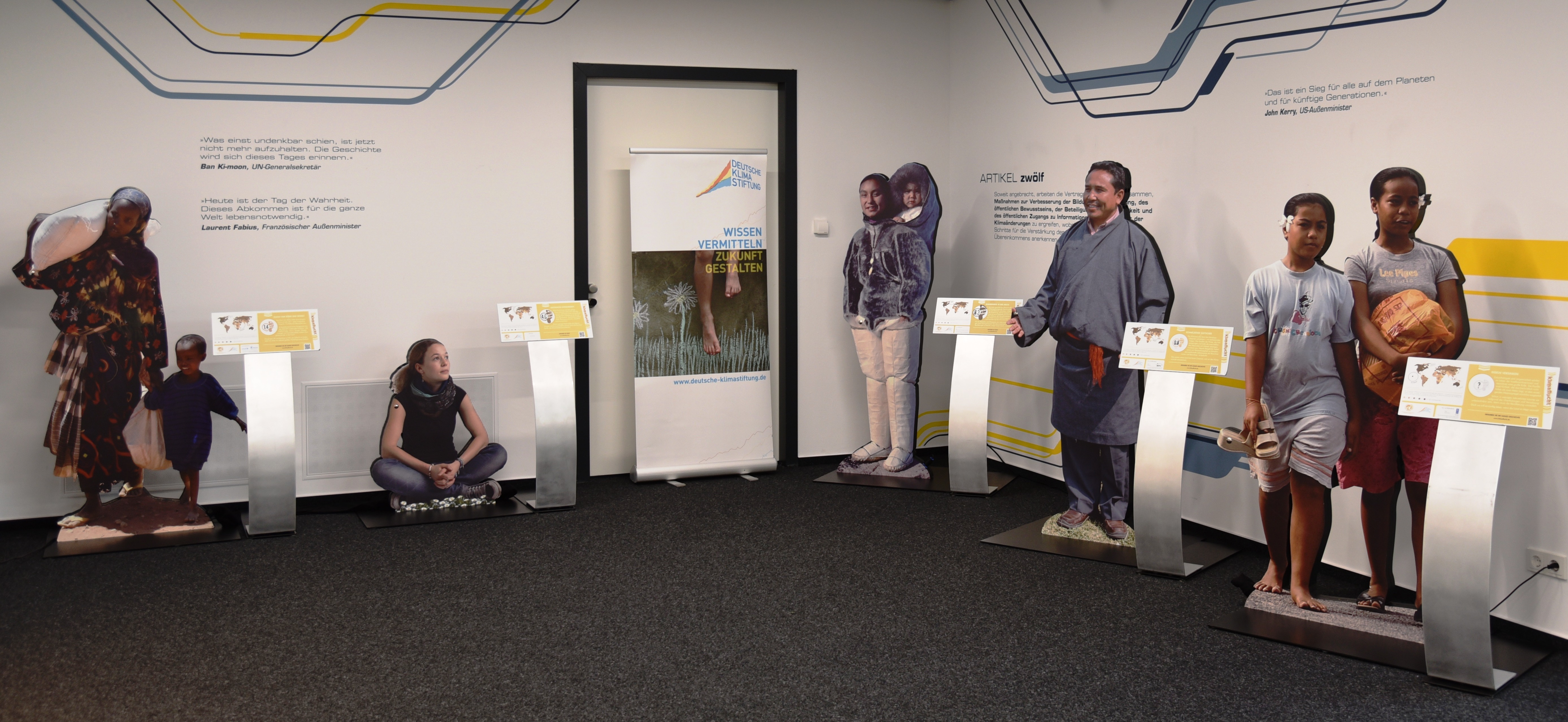 Aufnahme Teil der Klimafluchtausstellung. Lebensgroße pappfiguren verschiedener betroffener Personen stehen in einem Raum an der Wand, naben stehen Aufsteller mit Infotafeln.