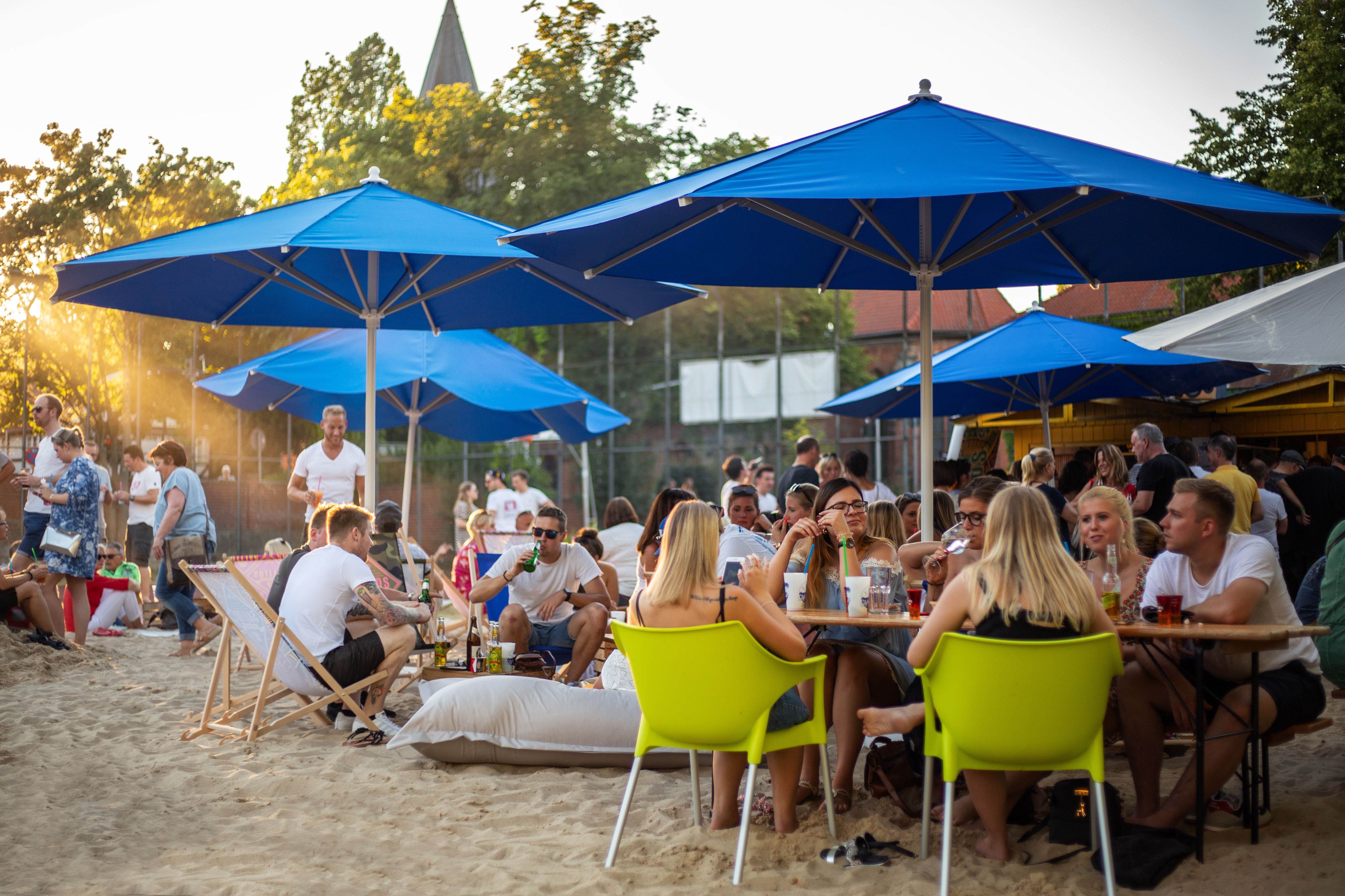 Blick auf den Betrieb des Citybeaches. Menschen sitzen an einem Sommerabend auf Liegestühlen unter Sonnenschirmen auf der Sandfläche vor dem Schloss und genießen Essen und Getränke.