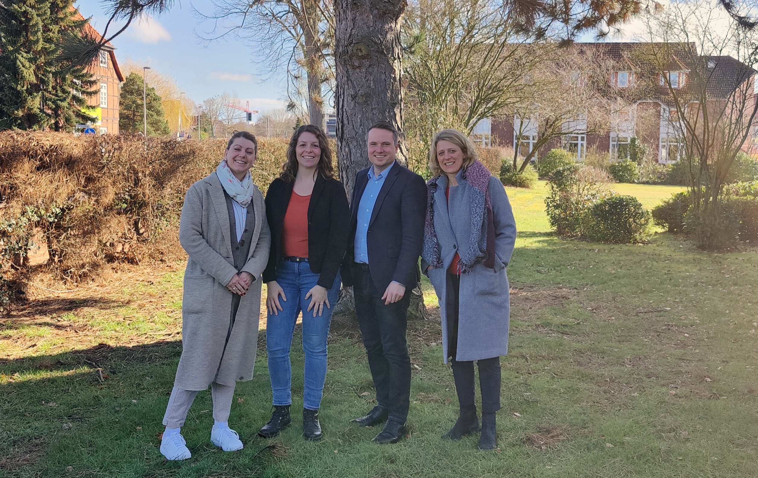 Vier Personen stehen vor einem Baum auf einer Wiese. Alle vier Lächeln freundlich in die Kamera.