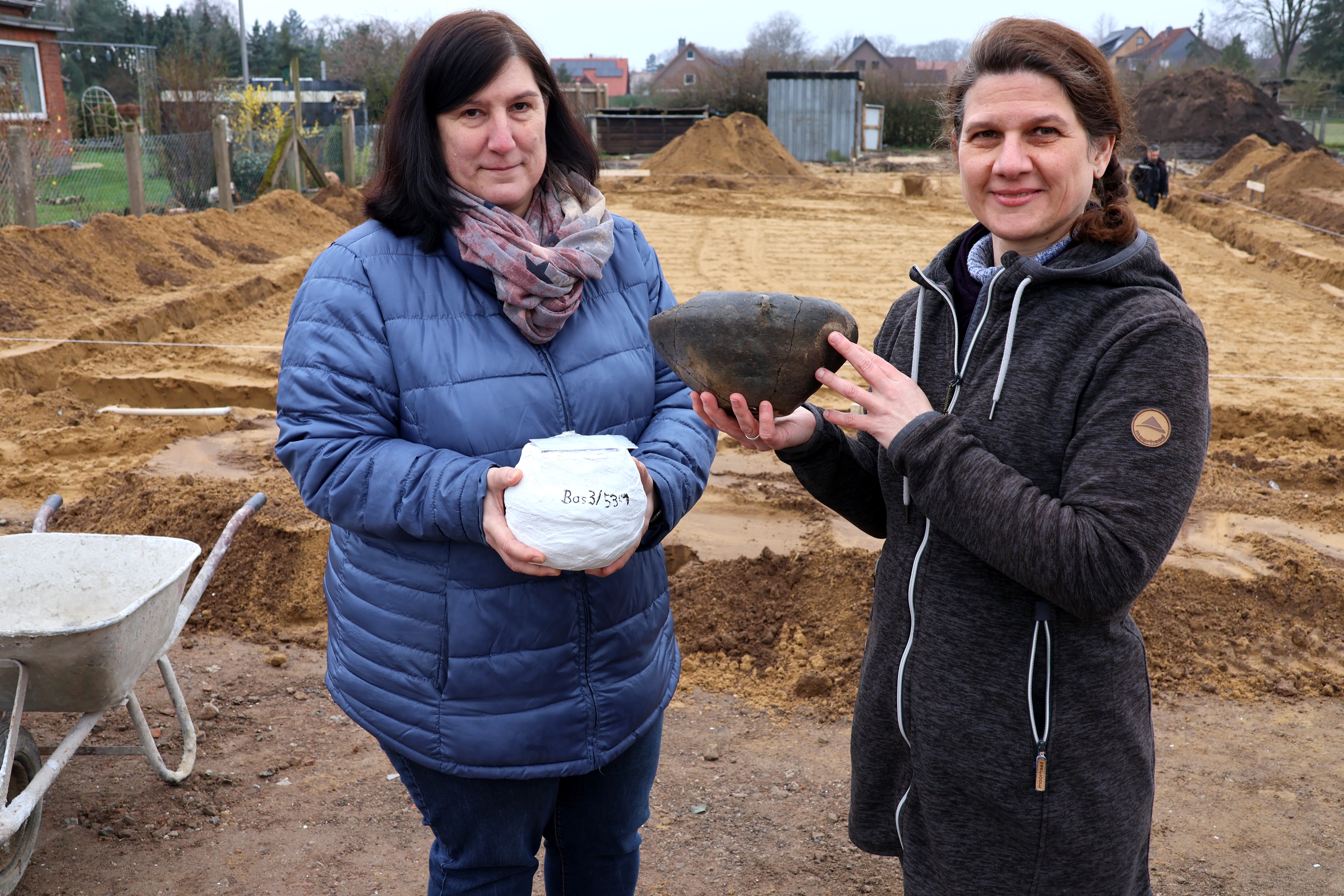 Die beiden Frauen stehen vor der Ausgrabunggstelle in Basse und halten je einen Fund in den Händen. Beide blicken in die Kamera.