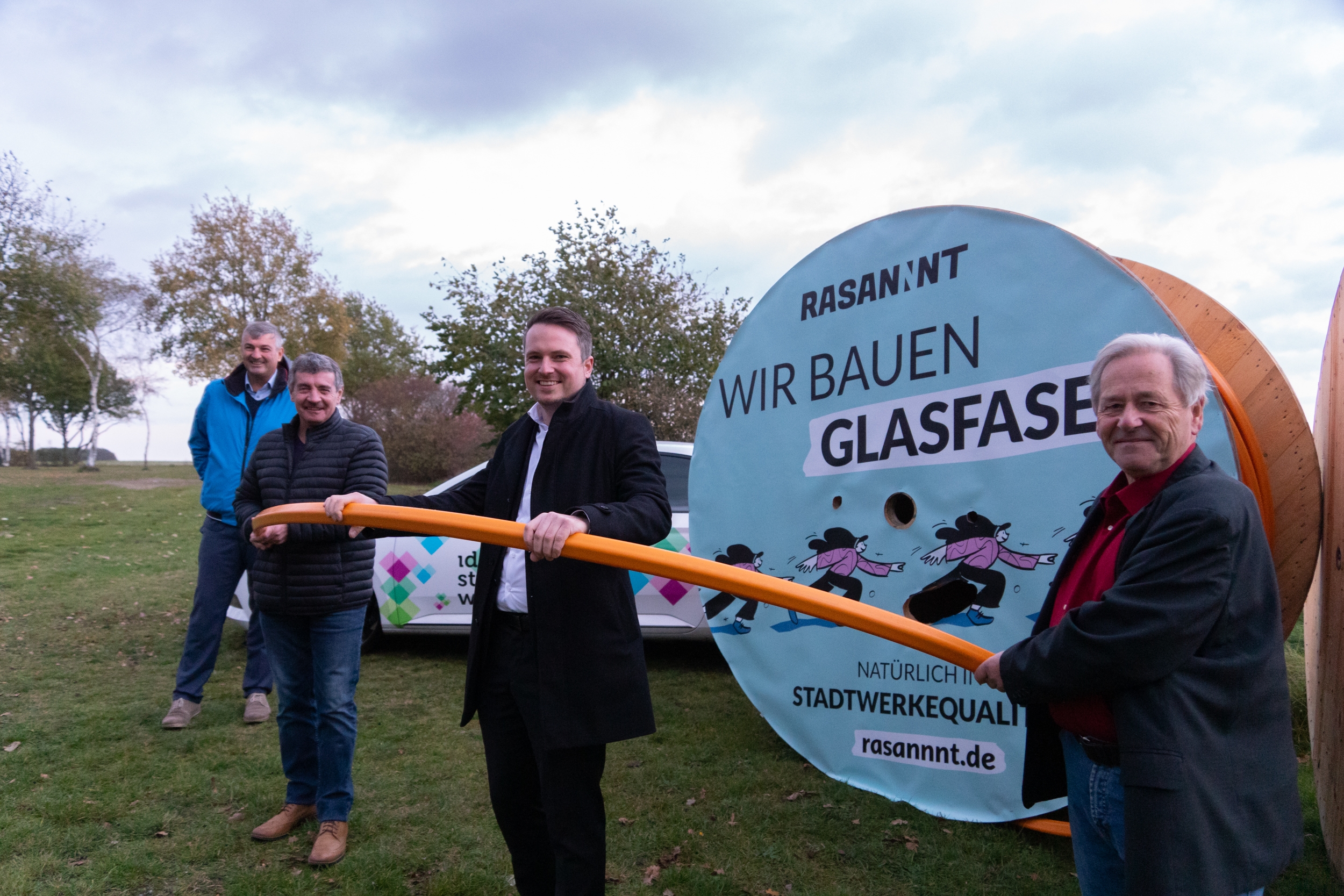 Vier Männer halten ein Glasfaserkabel. Sie stehen vor einer großen Kabeltrommel mit dem Schriftzug Rasannt. Wir bauen Glasfaser.