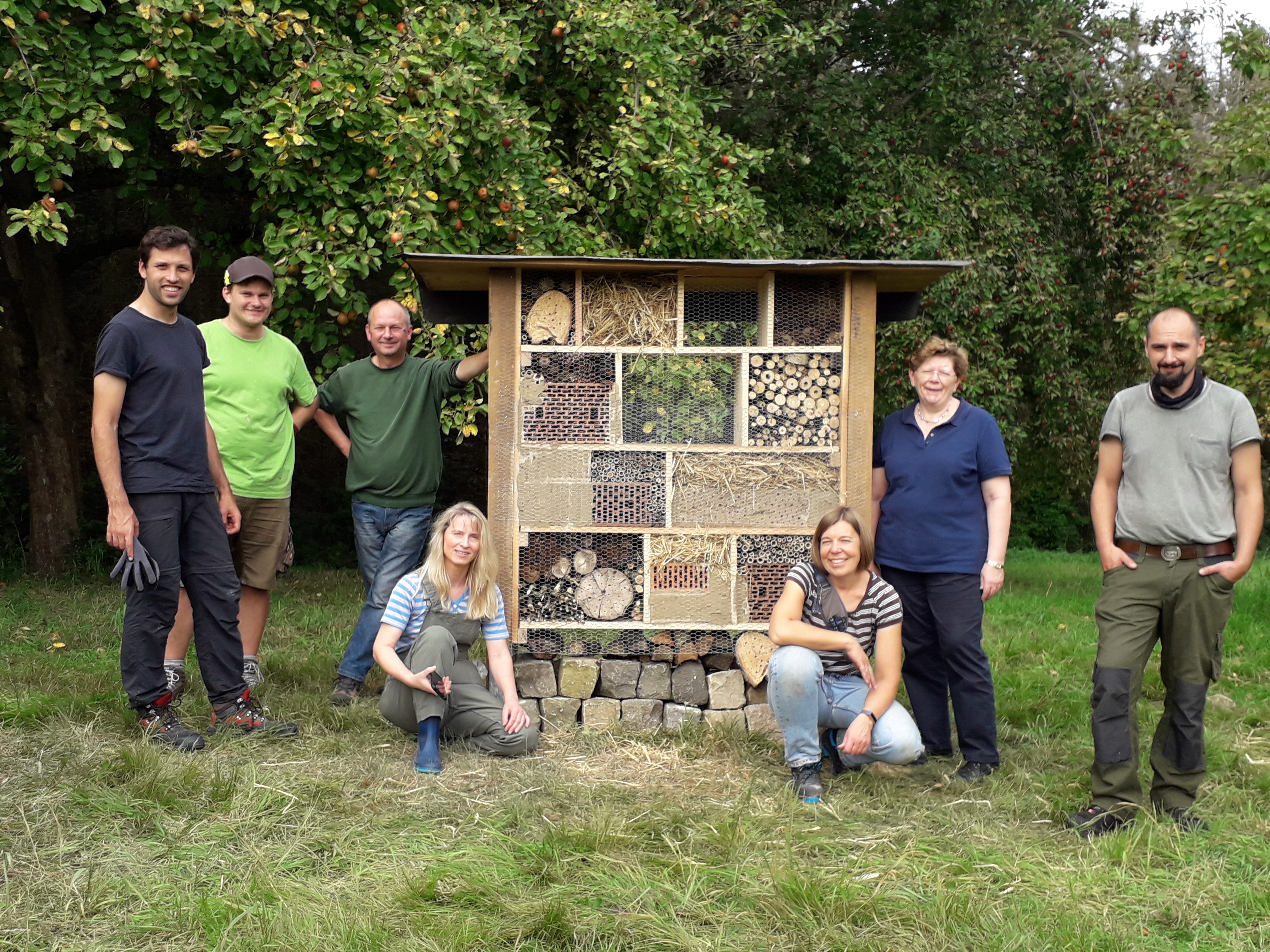 Ein Insektenhotel steht auf einer Wiese. Drum herum stehen und hocken die Mitarbeitenden des Fachdienstes Stadtgrün der Stadt Neustadt a. Rbge.