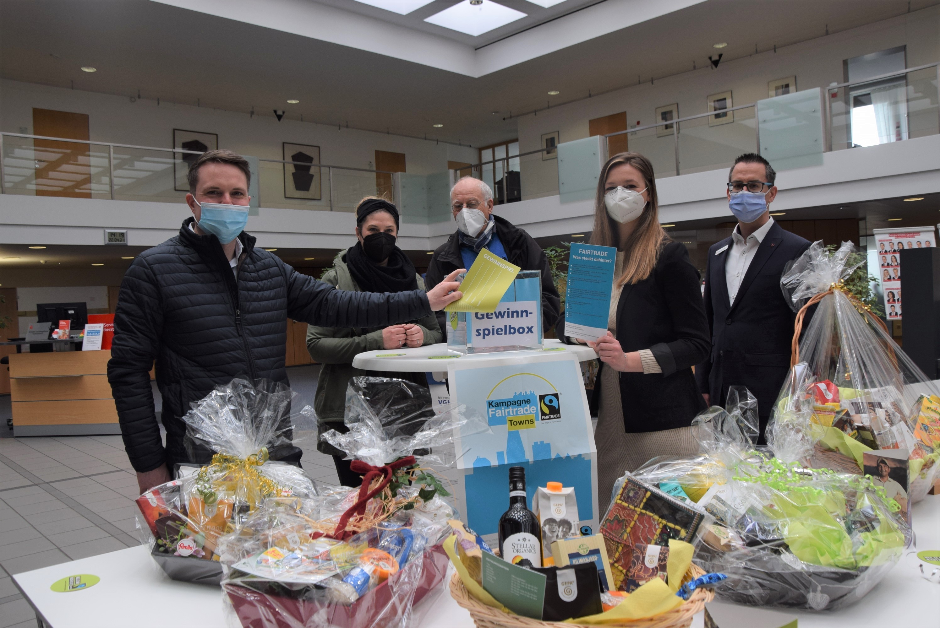 Fünf Personen mit Masken stehen hinter einem Tisch mit Präsentkörben. In der Mitte ist ein Stehtisch mit einer Gewinnspielbox. Zwei Personen halten die Quizflyer in die Kamera.