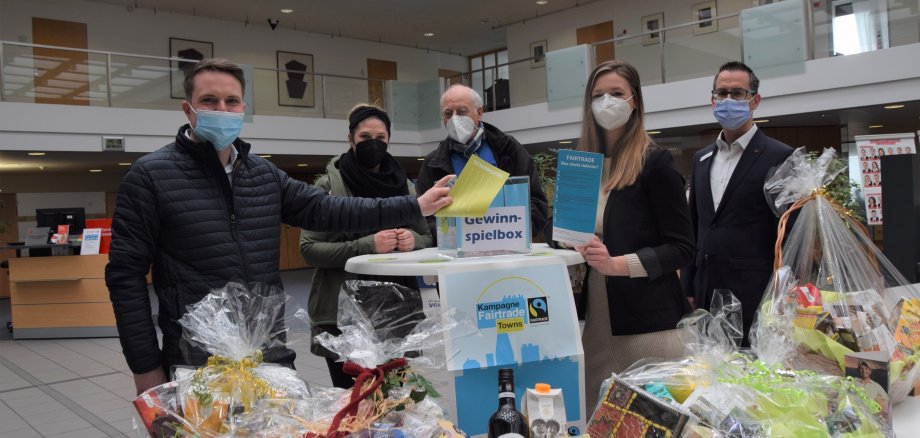 Neustadt wird Fairtrade Town Fünf Personen mit Masken stehen hinter einem Tisch mit Präsentkörben. In der Mitte ist ein Stehtisch mit einer Gewinnspielbox. Zwei Personen halten die Quizflyer in die Kamera.