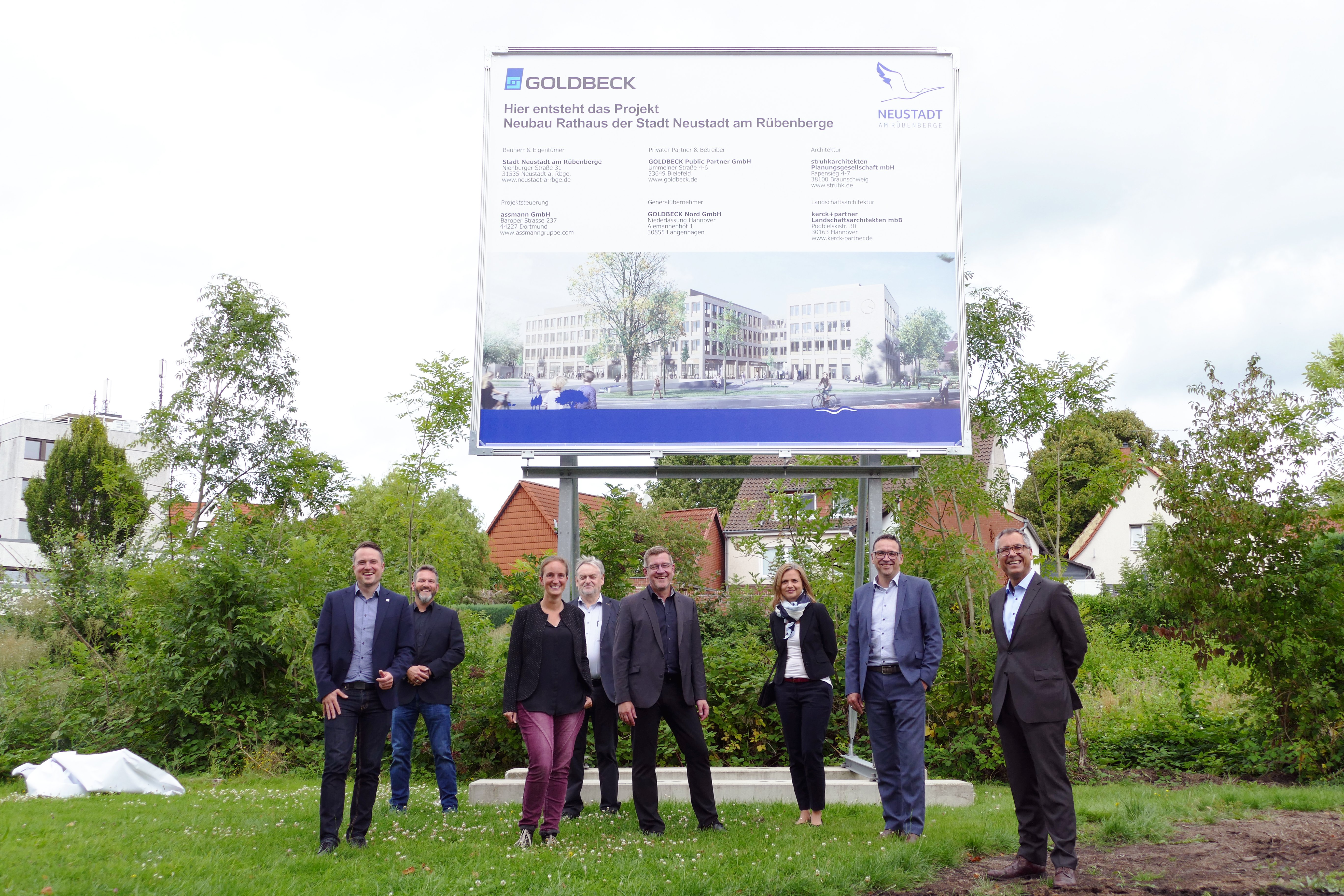 Acht Personen stehen auf einer Grünfläche vor einem rund vier meter hohem Baustellenschild. Dies zeigt den Rathausneubau und die beteiligten Firmen. 