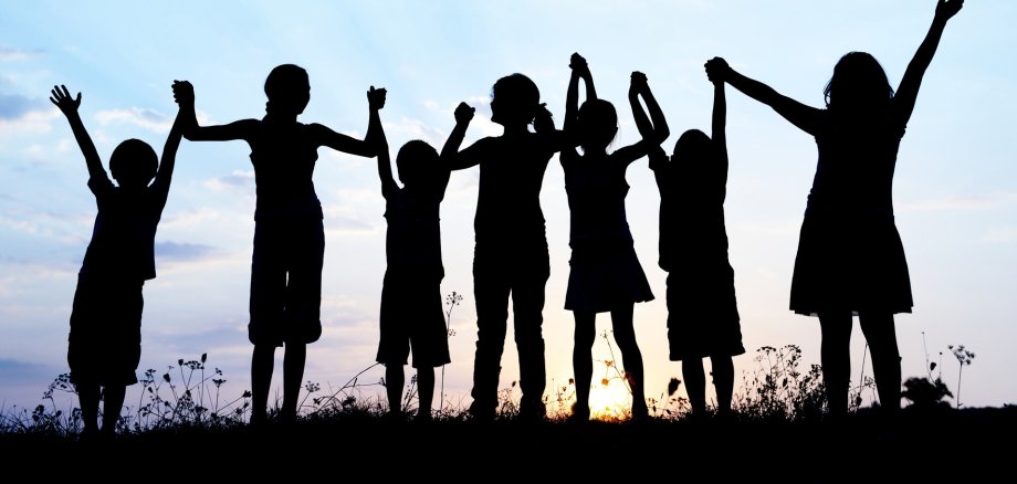 Symbolbild Kinder Freizeit sieben Kinder (nur als Schatten sichtbar) stehen auf einer Weise, halten sich an den Händen und strecken diese in die Höhe. Im Hintergrund geht die Sonne unter.
