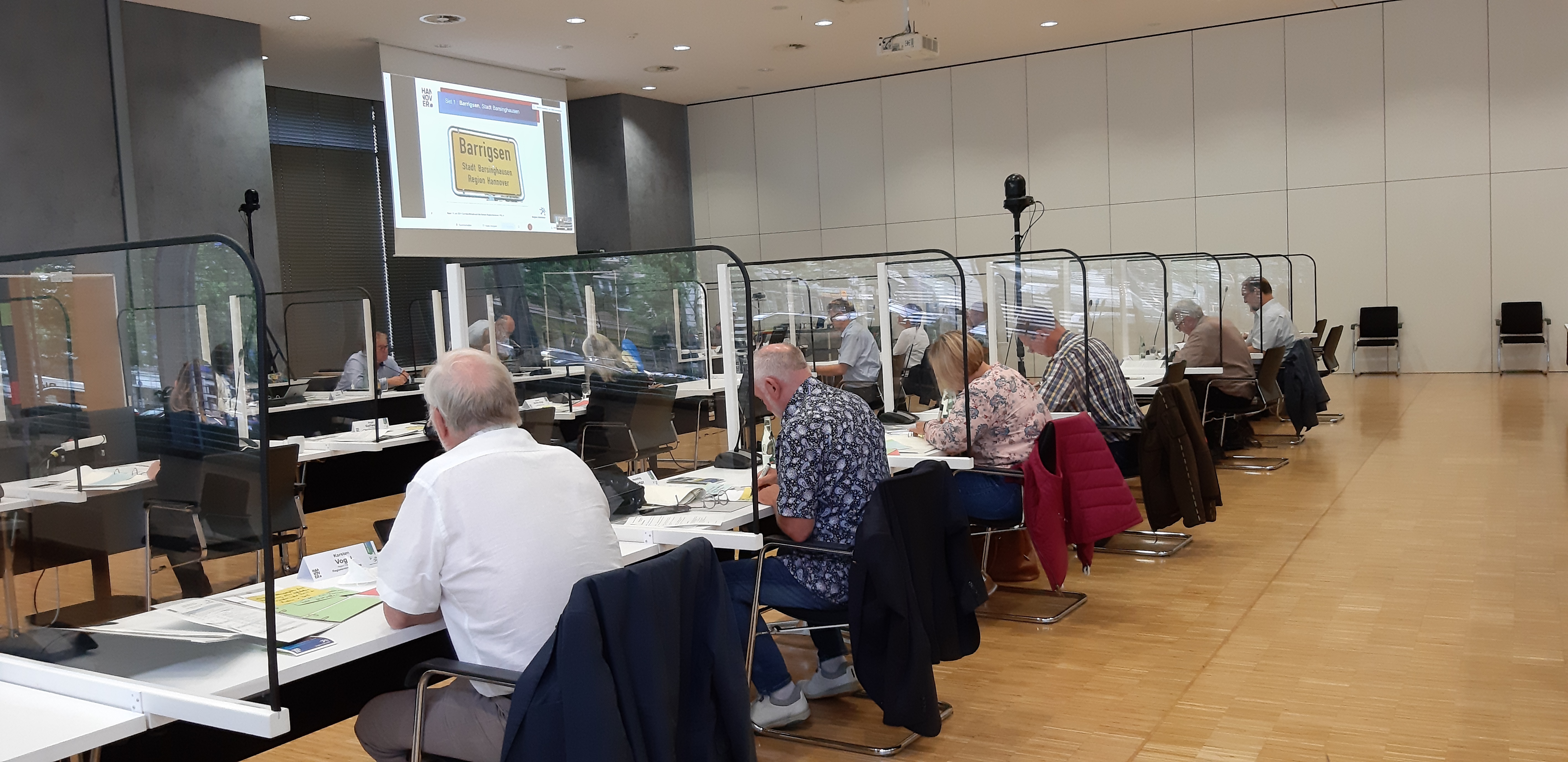 Blick in einen Konferenzsaal, dort sitzen Menschen an Tischen, getrennt von Plexiglas und bewerten die eingereichten Bewerbungen zum Wettbewerb.
