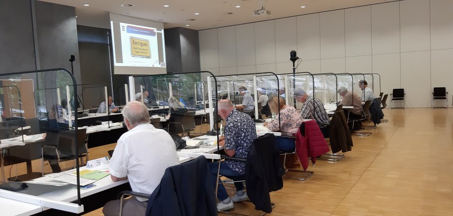 Blick in einen Konferenzsaal, dort sitzen Menschen an Tischen, getrennt von Plexiglas und bewerten die eingereichten Bewerbungen zum Wettbewerb.