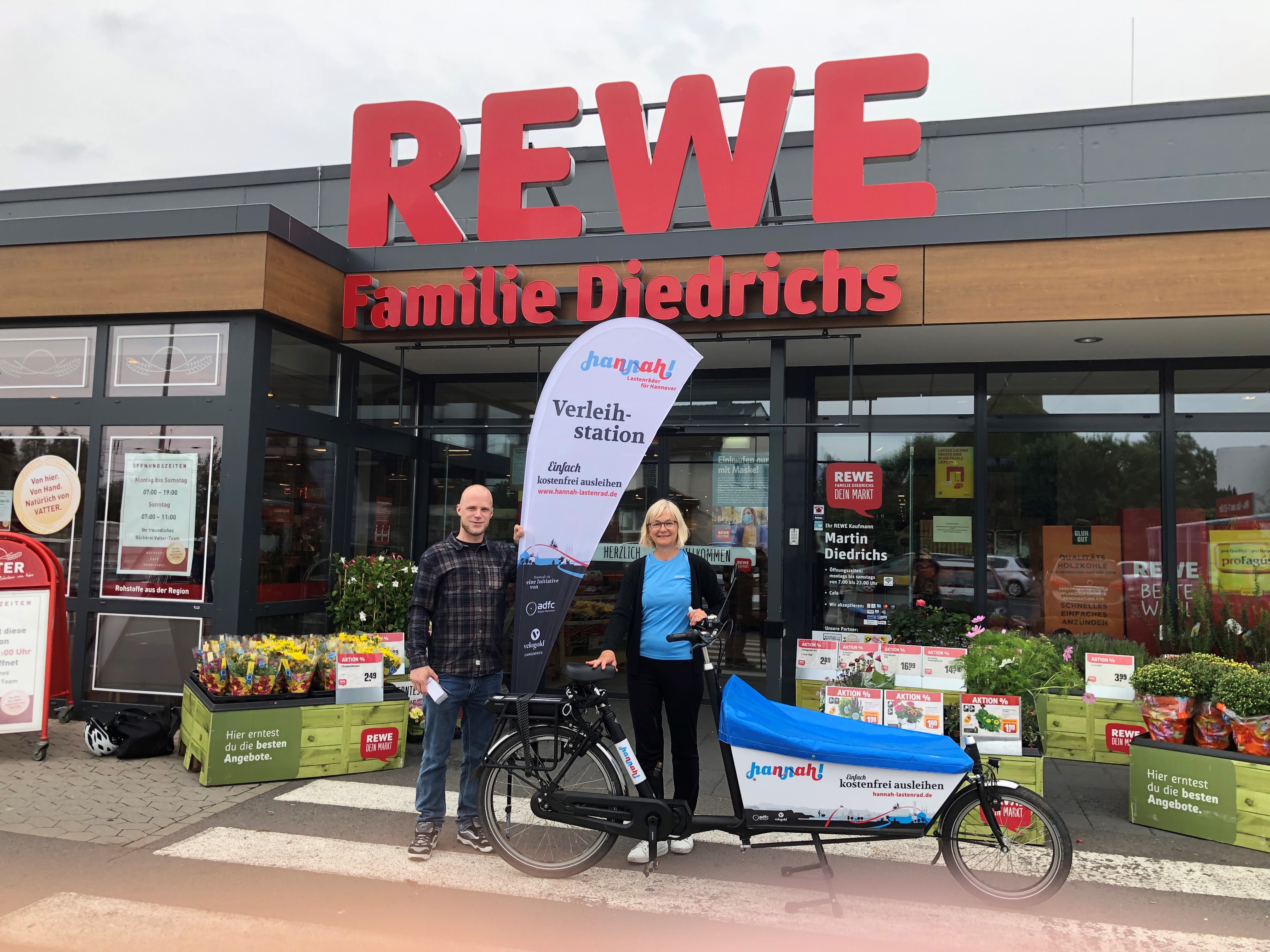 Zwei Personen stehen mit dem lastenrad Hannah und einer BEachflag vor dem Eingang des Rewew-Marktes an der Siemensstraße und lächeln in die Kamera