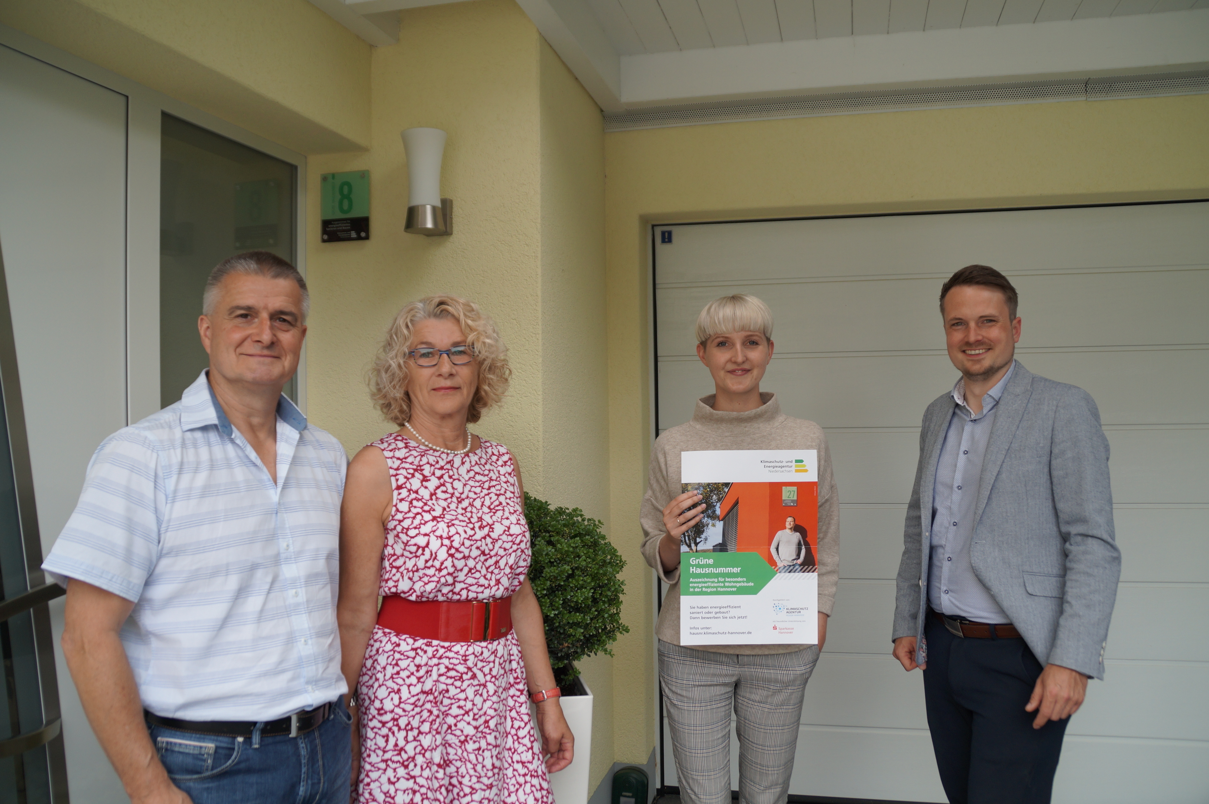 Vier Personen stehen vor einem Hauseingang, die zweite person von rechts hält das Zertifikat zur Grünen Hausnummer in den Händen, alle gucken in die Kamera.
