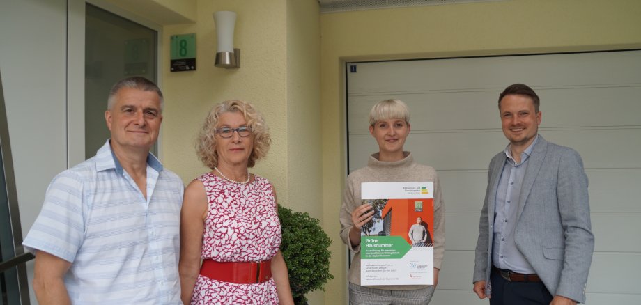 Pressefoto Grüne Hausnummer Vier Personen stehen vor einem Hauseingang, die zweite person von rechts hält das Zertifikat zur Grünen Hausnummer in den Händen, alle gucken in die Kamera.
