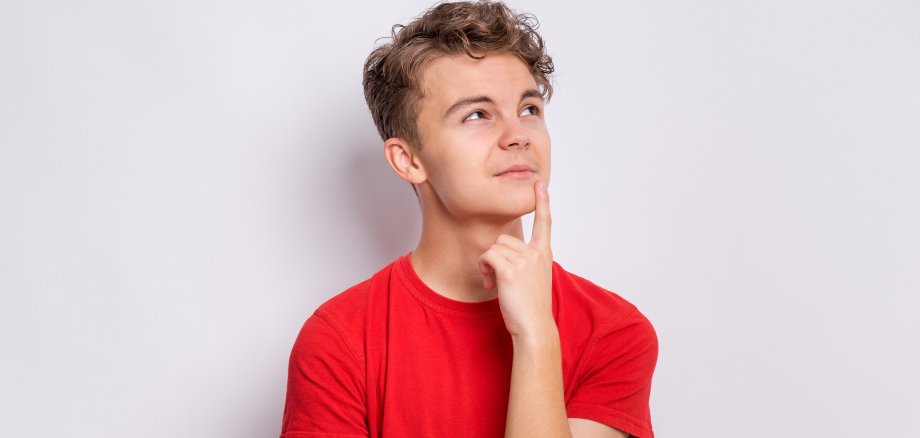 Symbolbild Frage/Umfrage Jugend Ein Jugendlicher im roten t-Shirt schaut nachdenklich nach oben rechts. Er steht vor hellem Hintergrund.