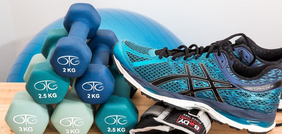 Symbolbild Sport. links ein Stapel mit kleinen Hanteln, rechts daneben ein Sportschuh. IM Hintergrund liegt ein Gymnastikball