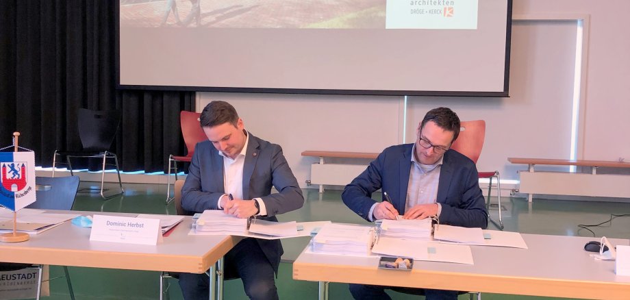 Zwei Männer sitzen an einem Tisch und unterschreiben Verträge. Im Hintergrund ist eine Leinwand, auf der das neue Rathaus zu sehen ist.