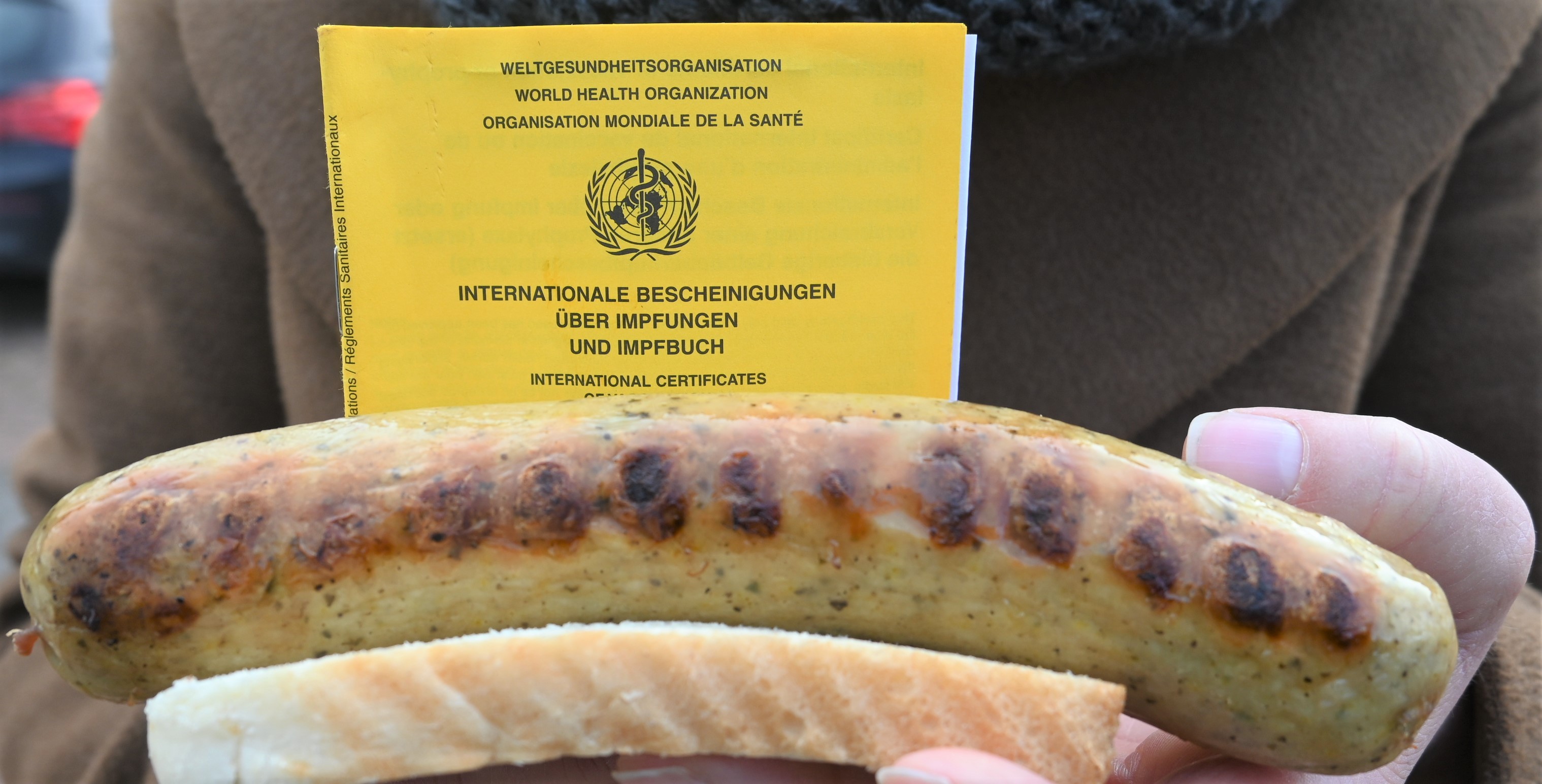 Eine Person hält eine Bratwurst in der Hand, dahinter ist ein gelber Impfpass zu sehen. 
