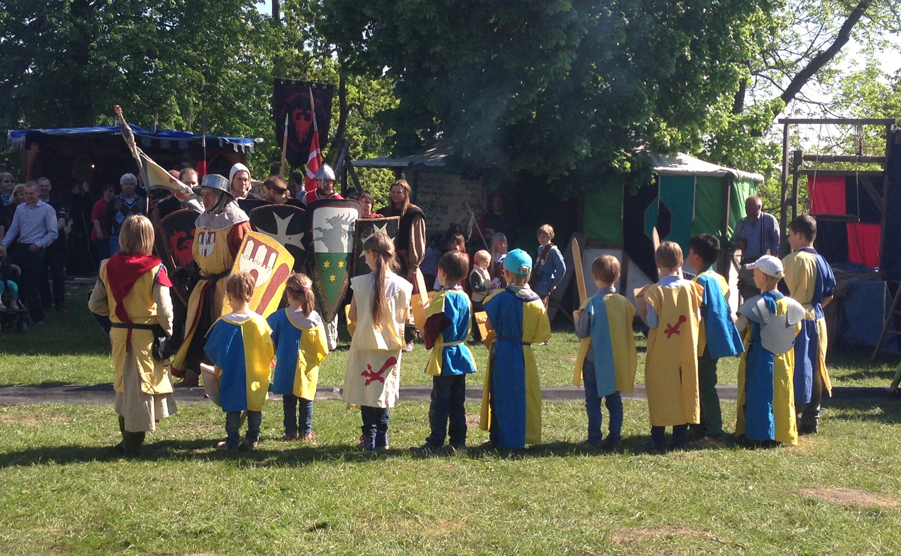Gruppe von Kindern in Knappenköstümen steht auf einer grünen Wiese. Im Hintergrund sind Mittealterzelte und Ritter zu sehen.