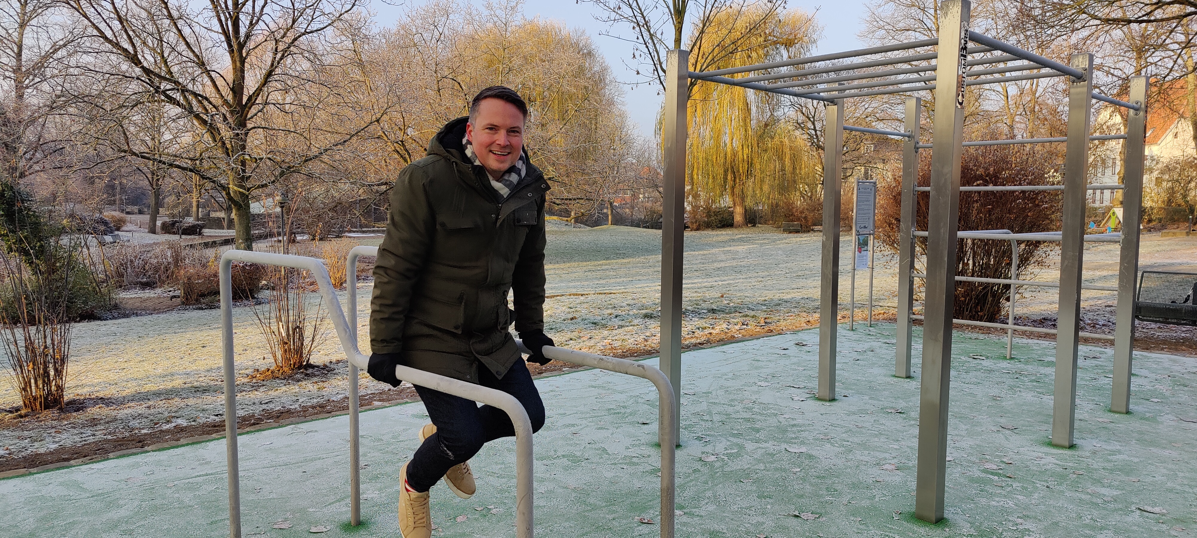 Ein junger Mann stemmt sich in Winterkleidung an zwei Metallstangen in die Luft. Im Hintergrund ist der Erichspark, sowie weitere Metallelemente der Calisthenics-Anlage zu sehen.