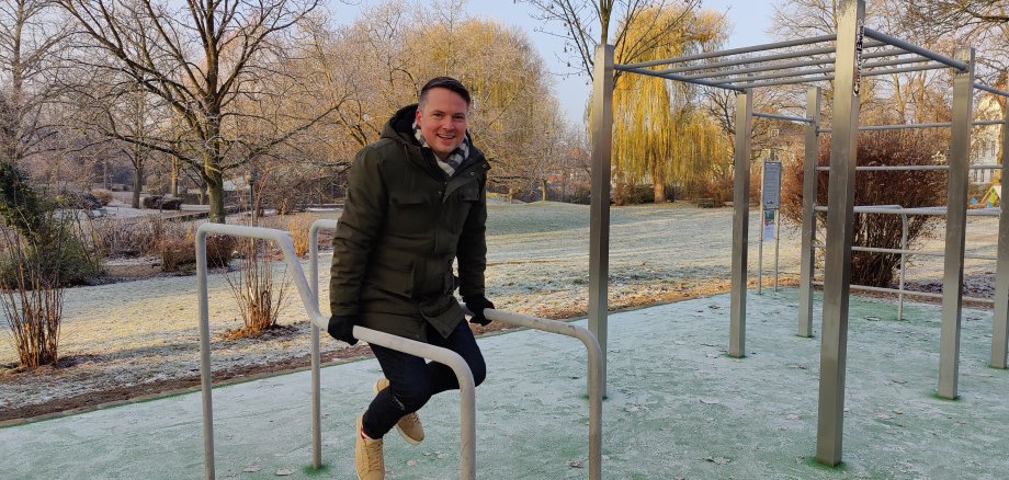 BGM Calisthenics Anlage Ein junger Mann stemmt sich in Winterkleidung an zwei Metallstangen in die Luft. Im Hintergrund ist der Erichspark, sowie weitere Metallelemente der Calisthenics-Anlage zu sehen.