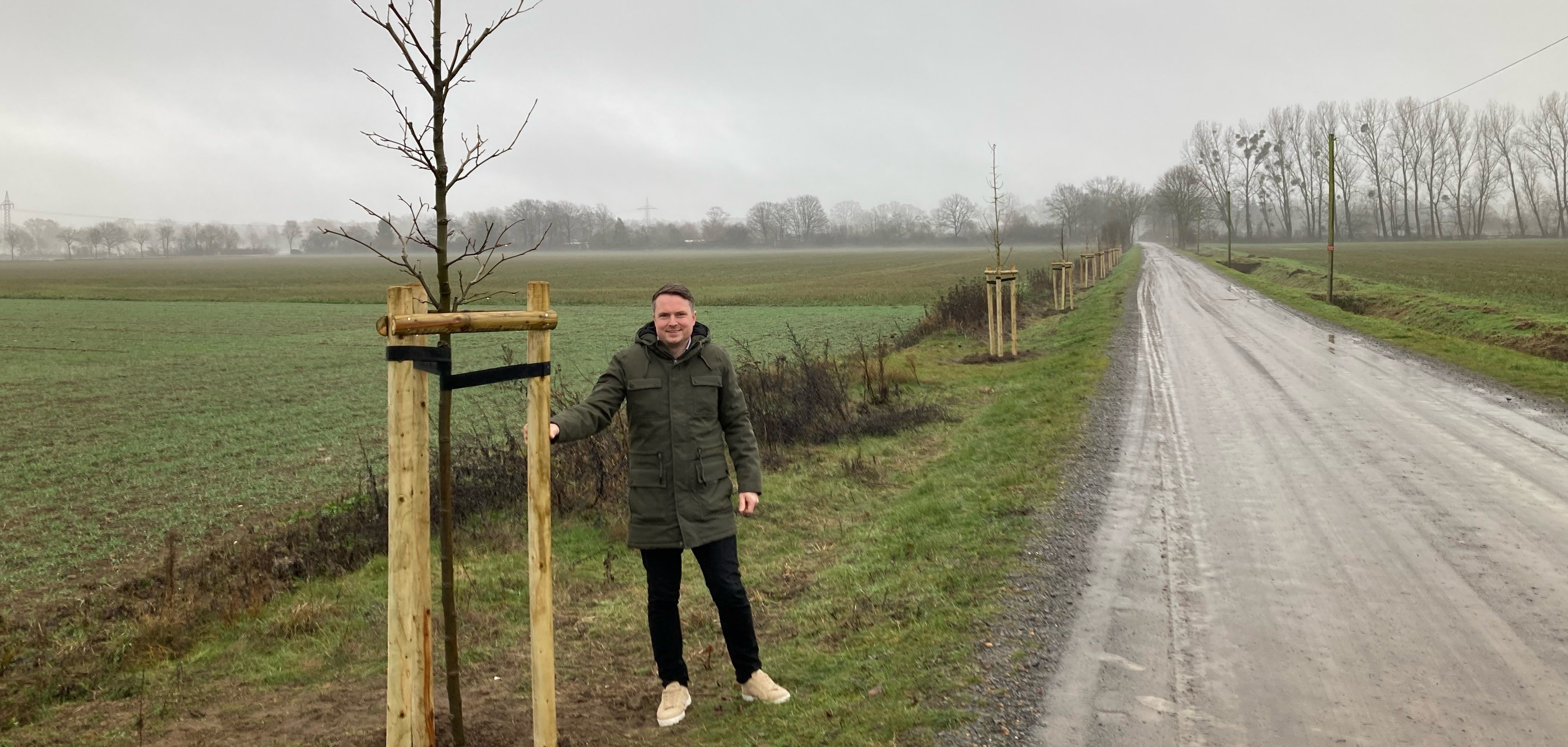 Bürgermeister herbst steht neben einem frisch geplfnzten Baum an einem Feldweg. Im Hintergrund sind Felder und weitere neu gepflanzte Bäume zu sehen. 
