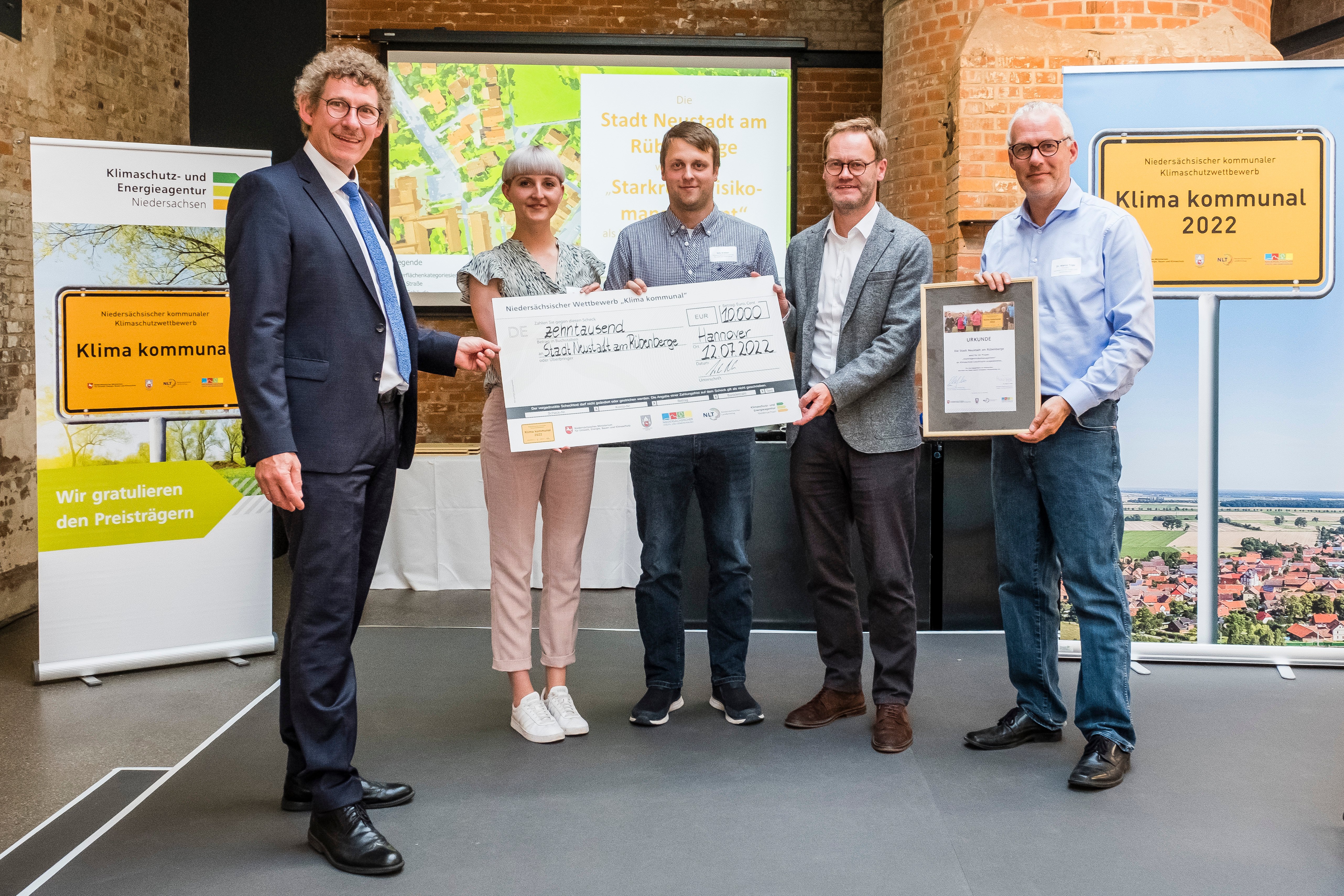 5 Personen stehen auf einer Bühne, die mittleren drei sind VertreterInnen der Stadt Neustadt, sie halten einen großen symbolischen Scheck in den Händen. Alle schauen in die Kamera.