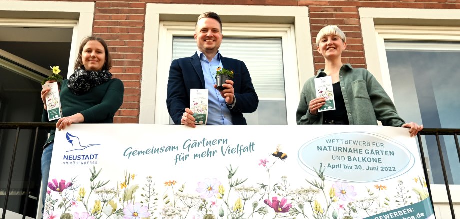 Pressefoto Gartenwettbewerb 2022 Drei personen stehen auf dem Balkon des Rathauses und halten Blumen und Samentüten in die Kamera. Am Balkongitter hängt ein Banner für den Garten- und Balkonwettbewerb.