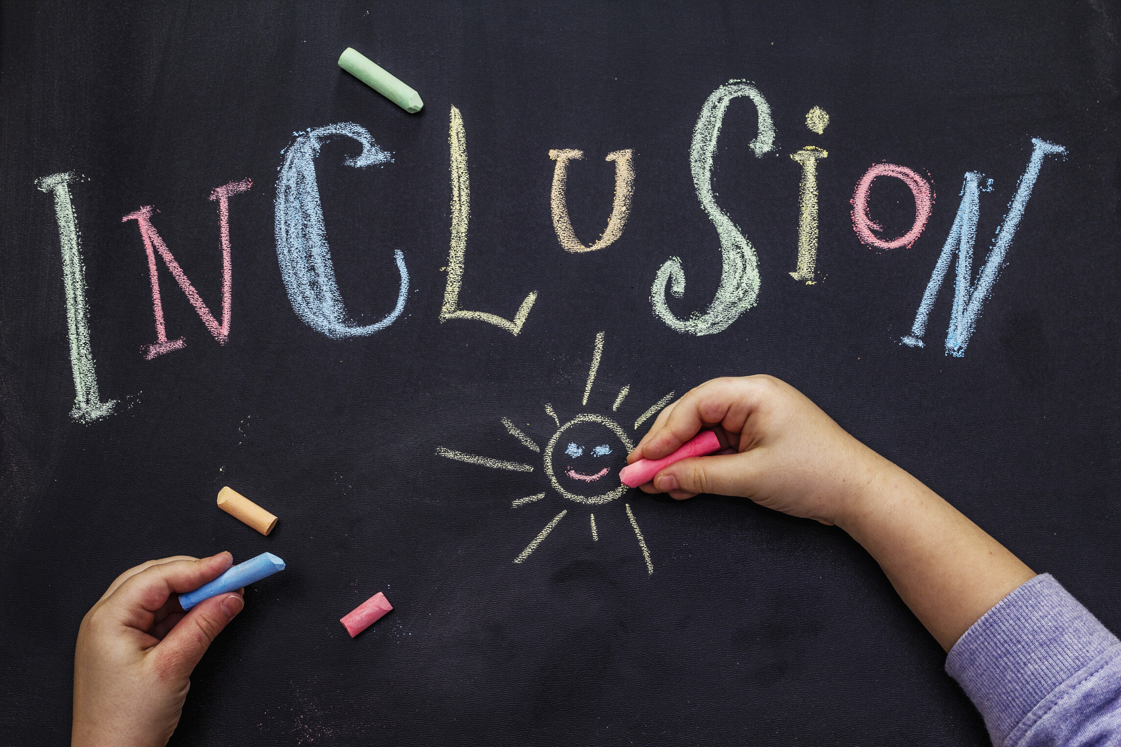 Auf einer schwarzen Tafels steht in bunten Kreisebuchstabden das Wort Inclusion. Darunter sind zwei Kinderhände zu sehen, die mit bunter Kreise eine Sonne malen.