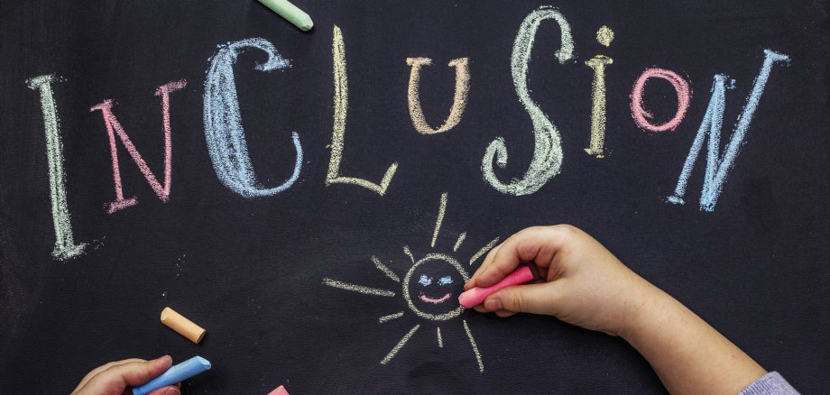 Symbolbild Inklusione Auf einer schwarzen Tafels steht in bunten Kreisebuchstabden das Wort Inclusion. Darunter sind zwei Kinderhände zu sehen, die mit bunter Kreise eine Sonne malen.